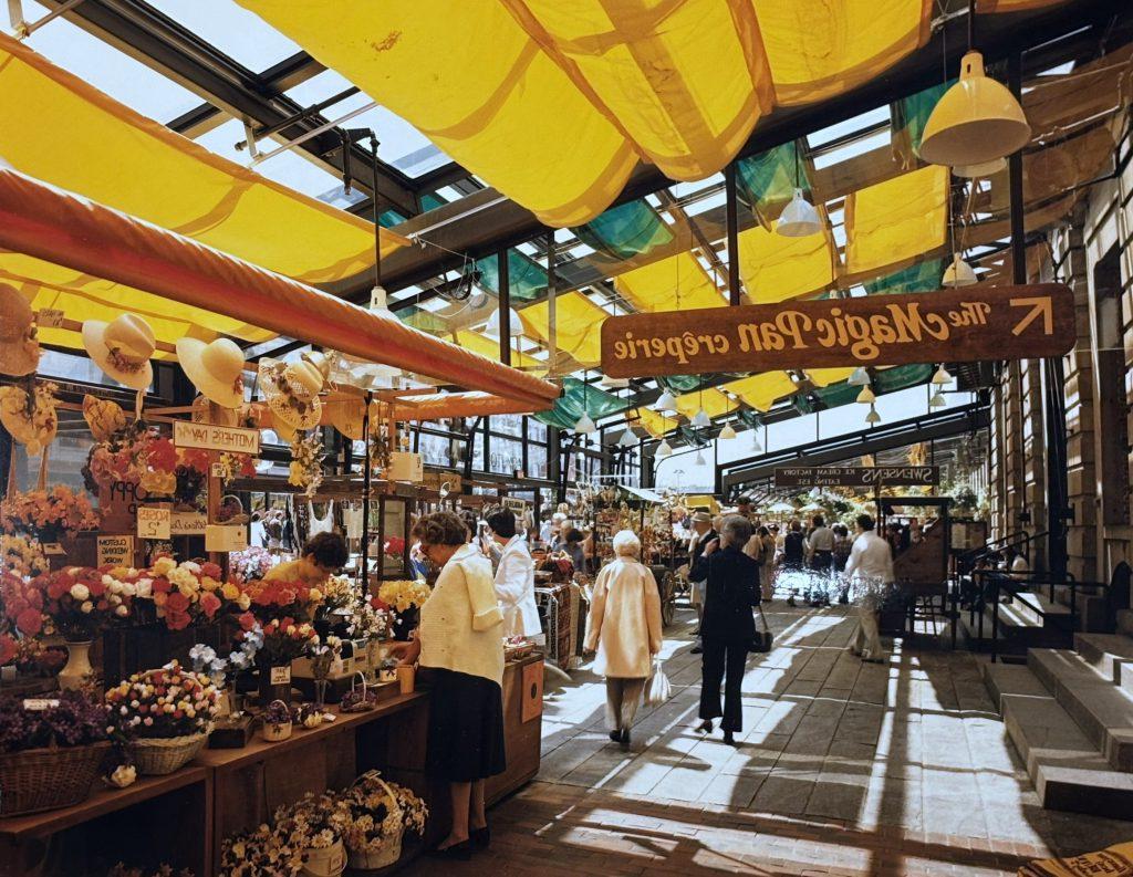 Shoppers browse market stalls selling flowers and other goods under colorful sun shades. A sign for "The Magic Pan Creperie" points away.